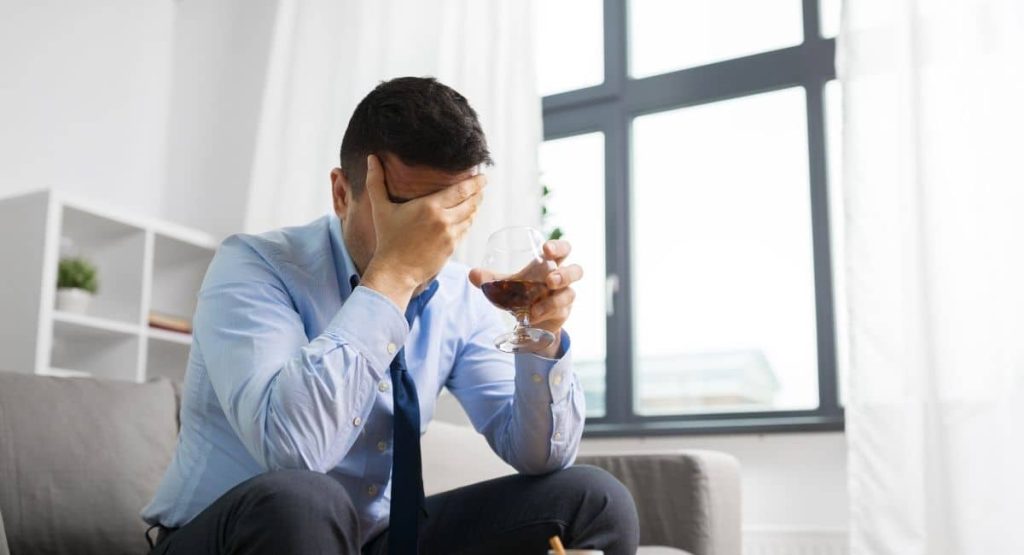 A man is head-rubbing while sitting on a couch and holding a glass of wine debating treating his alcoholism with ketamine for alcohol at Ketamine Therapy at Therapeutic Alternatives.