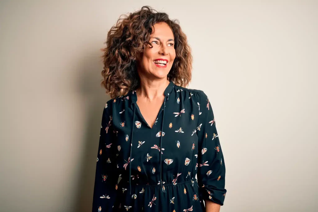 A smiling middle-aged woman with curly brown hair stands against a light neutral background from relief after using ketamine for perimenopause.
