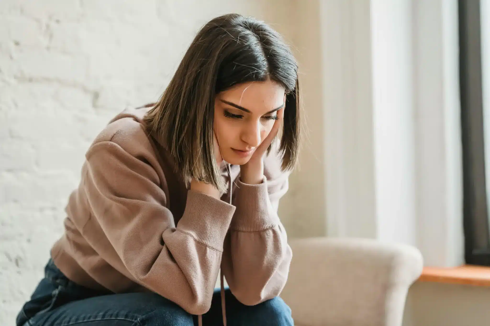 A woman sits indoors with her head lowered and hands near her neck as she contemplates ketamine for seasonal depression treatment at Ketamine Therapy at Therapeutic Alternatives.